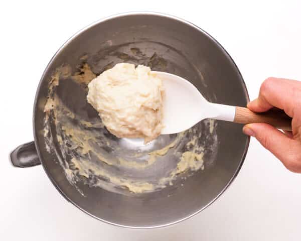 A hand holds a spatula with flour paste, hovering over a mixing bowl.