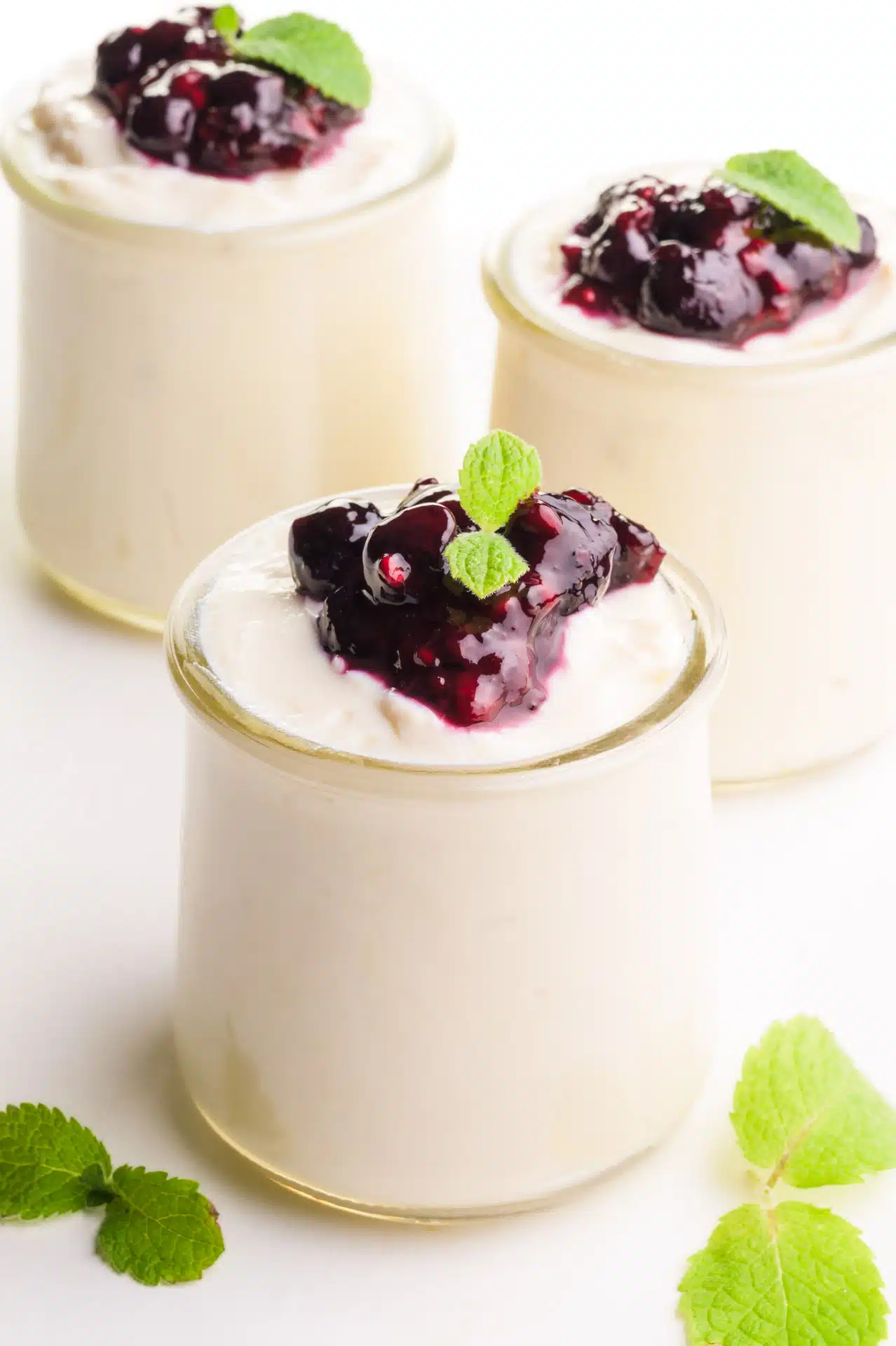 Three containers of soy yogurt have a fruit topping and mint leaves. There are mint leaves on the table too. 