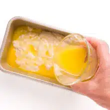 A hand pours melted butter over raw bread dough in a loaf pan.