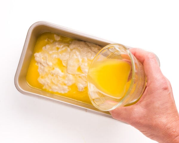 A hand pours melted butter over raw bread dough in a loaf pan.