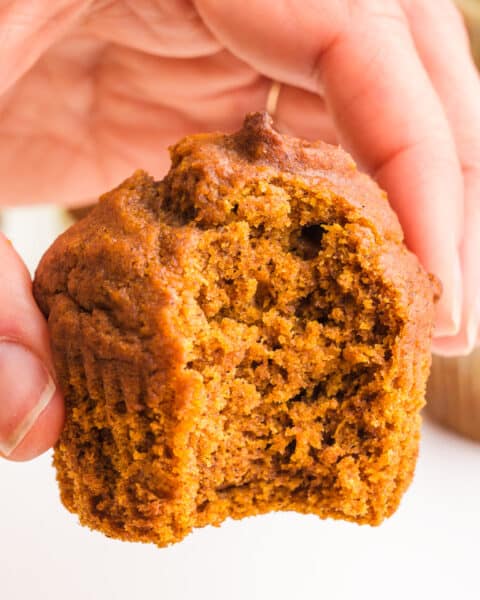 A hand holds a plant-based pumpkin muffin with a bite taken out.