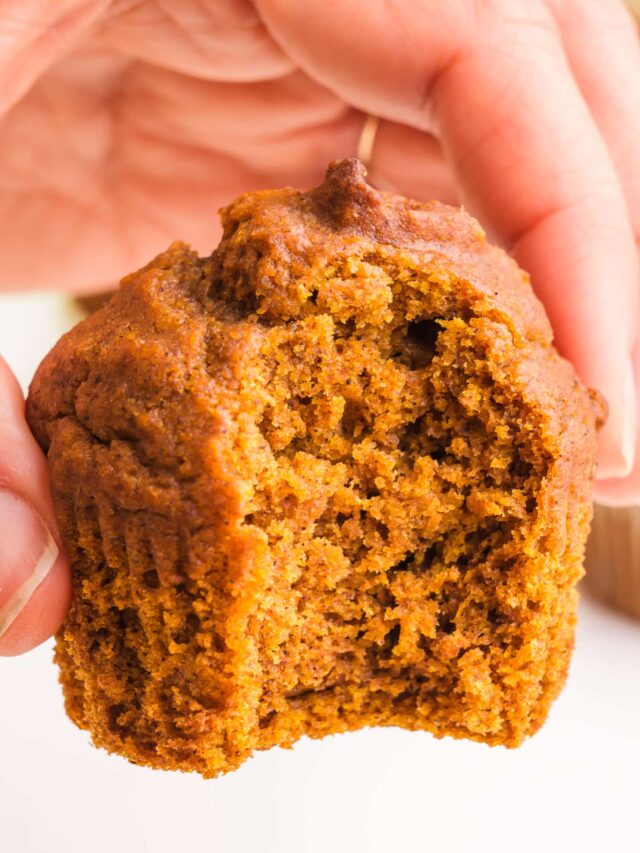 A hand holds a plant-based pumpkin muffin with a bite taken out.