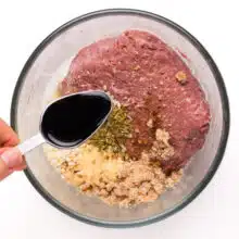 An artful pour of vegan Worcestershire sauce into a bowl filled with a tantalizing mix of Impossible Meat, breadcrumbs, and a medley of flavorful ingredients. The hands-on preparation captures the essence of crafting delicious and plant-powered Impossible Meatballs.