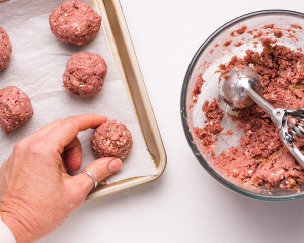 Hands at work in the kitchen as uncooked Impossible Burger meatballs are carefully placed on a baking sheet. A bowl filled with the flavorful meatball mixture and a convenient dispenser sit nearby, highlighting the easy and enjoyable process of preparing these plant-based delights.