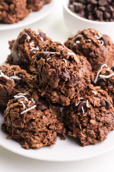 A stack of protein no-bake cookies sit on a plate. There are more cookies in the background along with a bowl of chocolate chips.
