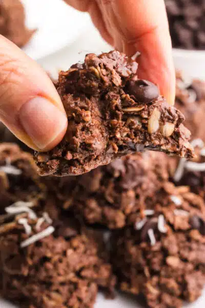 A hand holds a no-bake high protein cookie with a bite taken out. It hovers over a plate with more of the cookies.