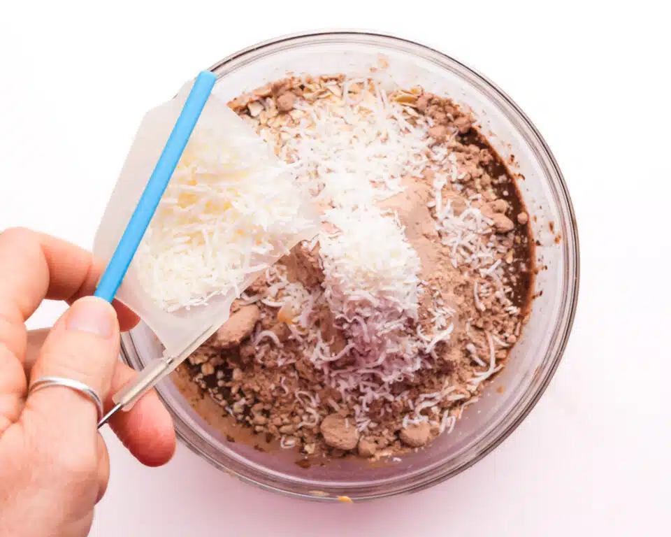 Coconut flakes are being poured into a mixing bowl with other ingredients, including chocolate protein powder.