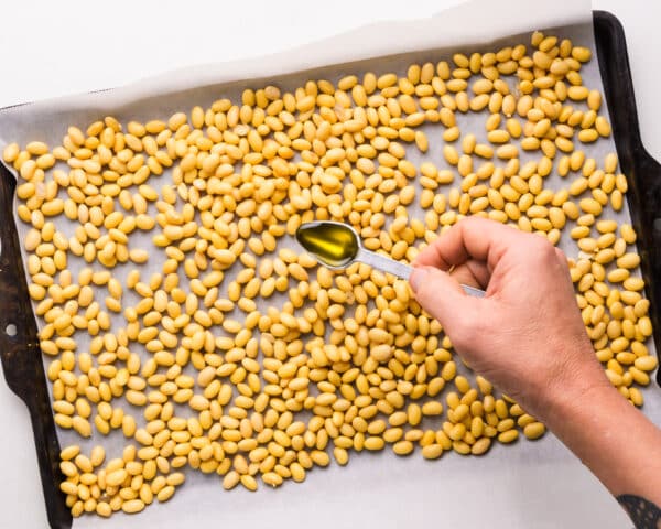 A hand holds a spoon, drizzling olive oil over soybeans in a pan.