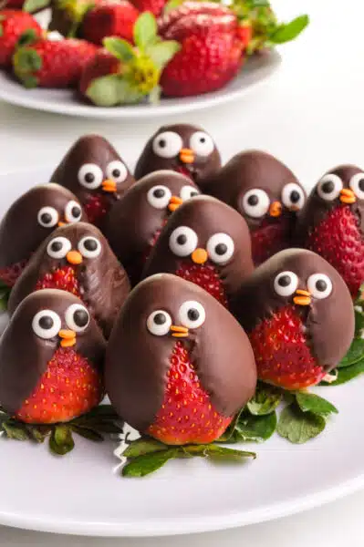 A plate full of chocolate-covered strawberry penguins sit on a plate. There are more strawberries in the background.