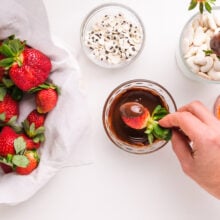 A hand holds a strawberry, dipping it in melted chocolate. There is a plate of strawberries and bowls of candy eyes next to the melted chocolate.