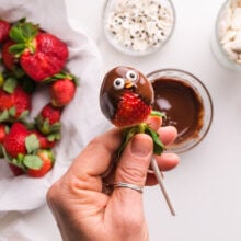 A hand holds a stick with a chocolate dipped strawberry with two candy eyes on it. There is a plate of strawberries and bowls of candies below it.