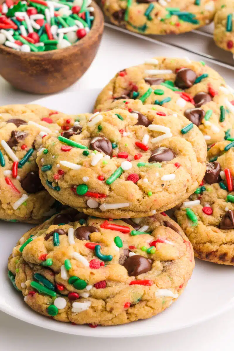 A plate holds Christmas cookies with chocolate chips. There's a bowl of sprinkles and more cookies in the background.