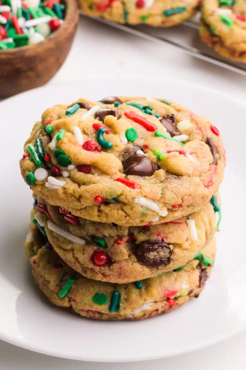 A stack of chocolate chip Christmas cookies sit on a plate. There are more cookies and a bowl of sprinkles in the background.