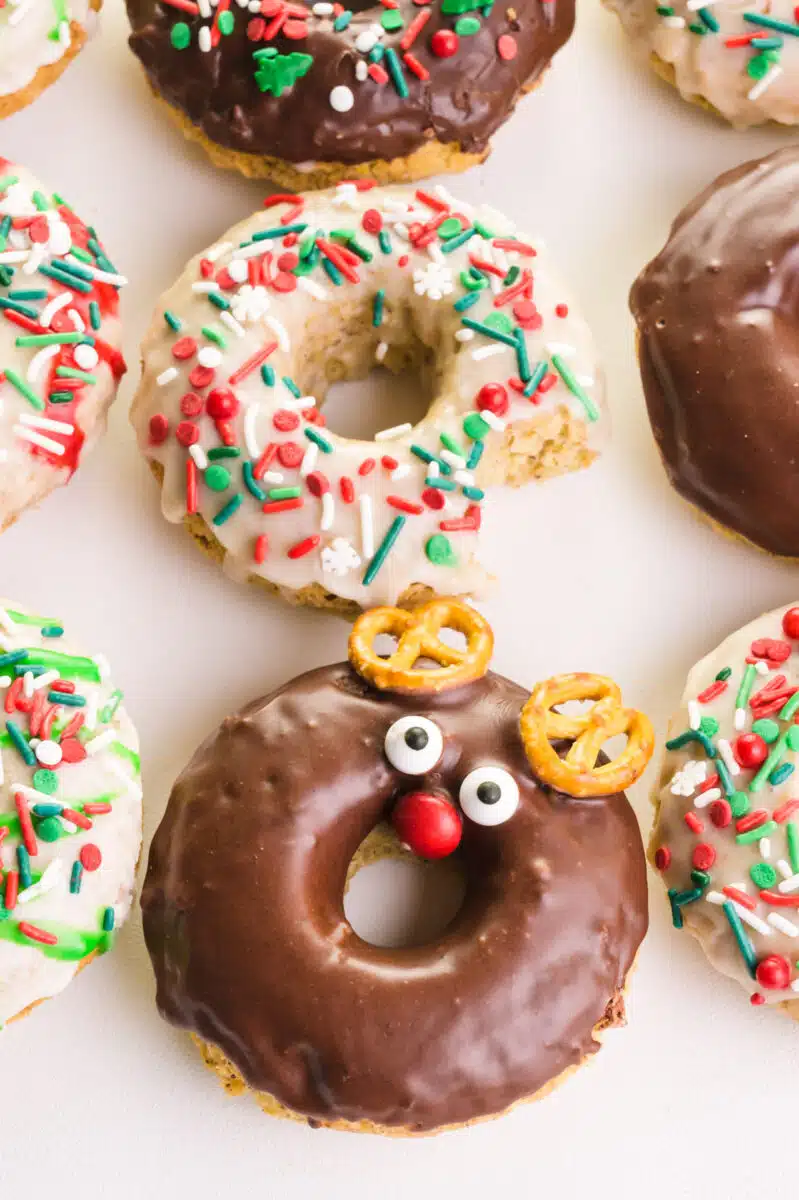 Looking down on several baked donuts with Christmas toppings. One has a bite taken out and one is decorated like a reindeer.