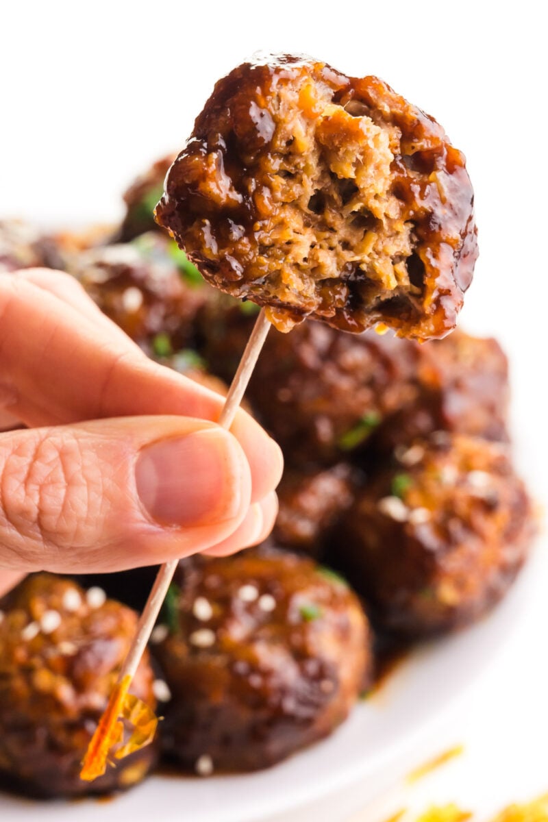 A hand holds a vegan jelly meatball on a toothpick. There is a bite out of the meatball and more meatballs behind it.