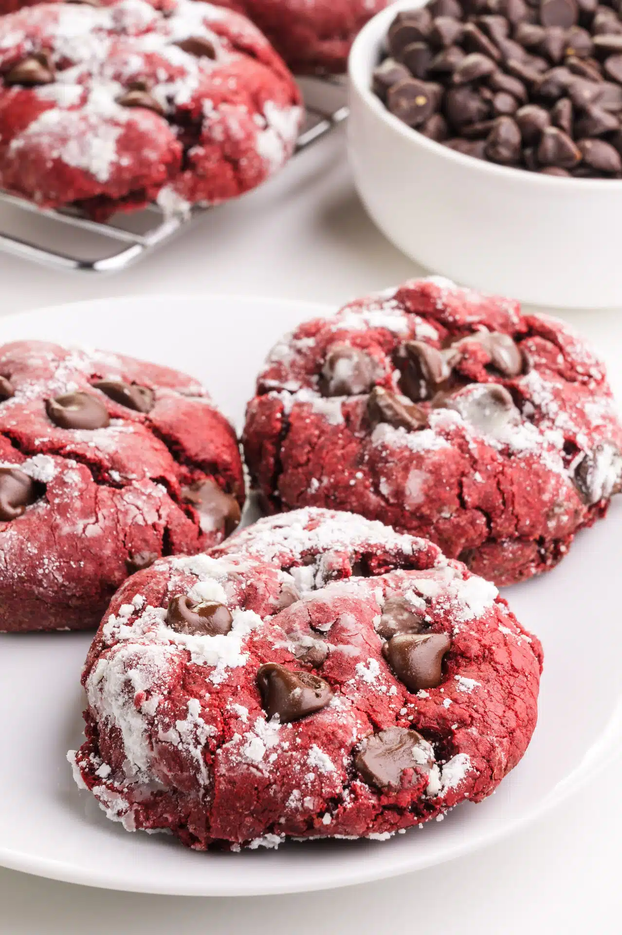 Red velvet chocolate chip cookies are on a plate. There's a bowl of chocolate chips and more cookies in the background.