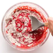 A hand holds a spatula stirring red velvet batter in a mixing bowl.
