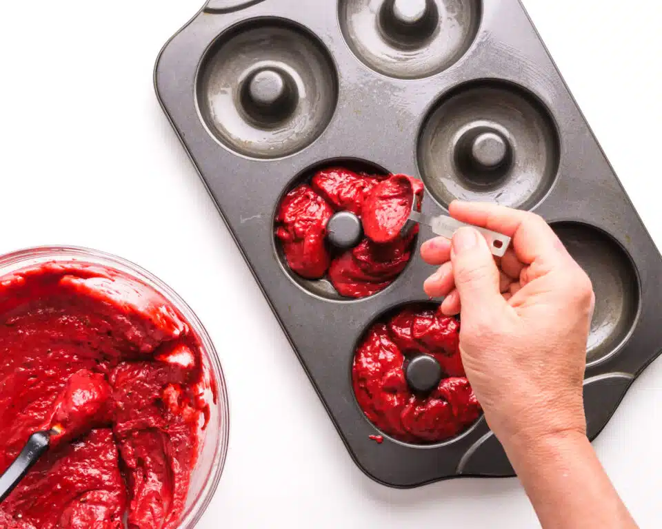 A hand distributes red velvet batter into a donut baking pan.