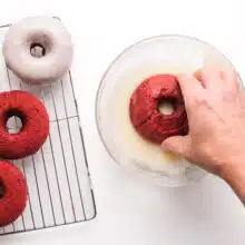 A hand dips a baked red velvet doughnut into icing. There is a wire rack next to it with more donuts.