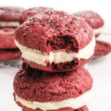 A stack of vegan red velvet whoopee pies, shows the top one with a bite taken out. There are more of the treats in the background.