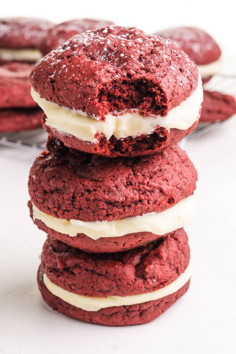 A stack of vegan red velvet whoopee pies, shows the top one with a bite taken out. There are more of the treats in the background.