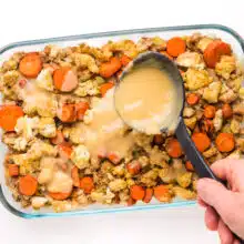 A hand holds a ladle, drizzling gravy over the top of a casserole with carrots and tofu.