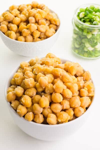 Two bowls of chickpeas sit on a counter next to a jar of chopped green onions.