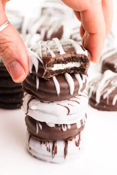A stack of vegan chocolate-covered Oreos shows the top one with a bite taken out. A hand reaches for the top cookie.