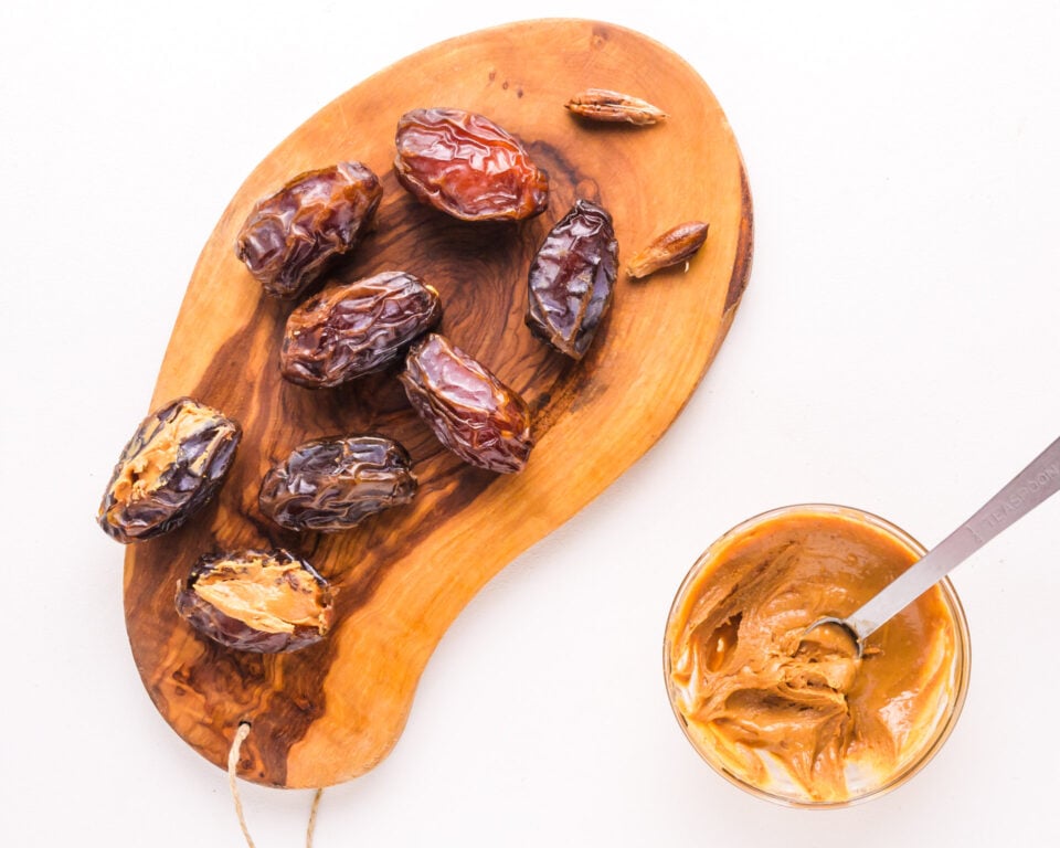 Looking down on Medjool dates on a cutting board sitting next to a bowl of peanut butter with a spoon in it.
