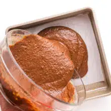 A step-by-step process shot capturing the pouring of chocolate cake batter into a square cake pan lined with parchment paper. The image illustrates the initial stage of creating the delectable whole wheat healthy chocolate cake, with the glossy batter smoothly filling the pan.