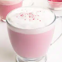 A hot mug of pink matcha latte has another mug and matcha powder in the background.