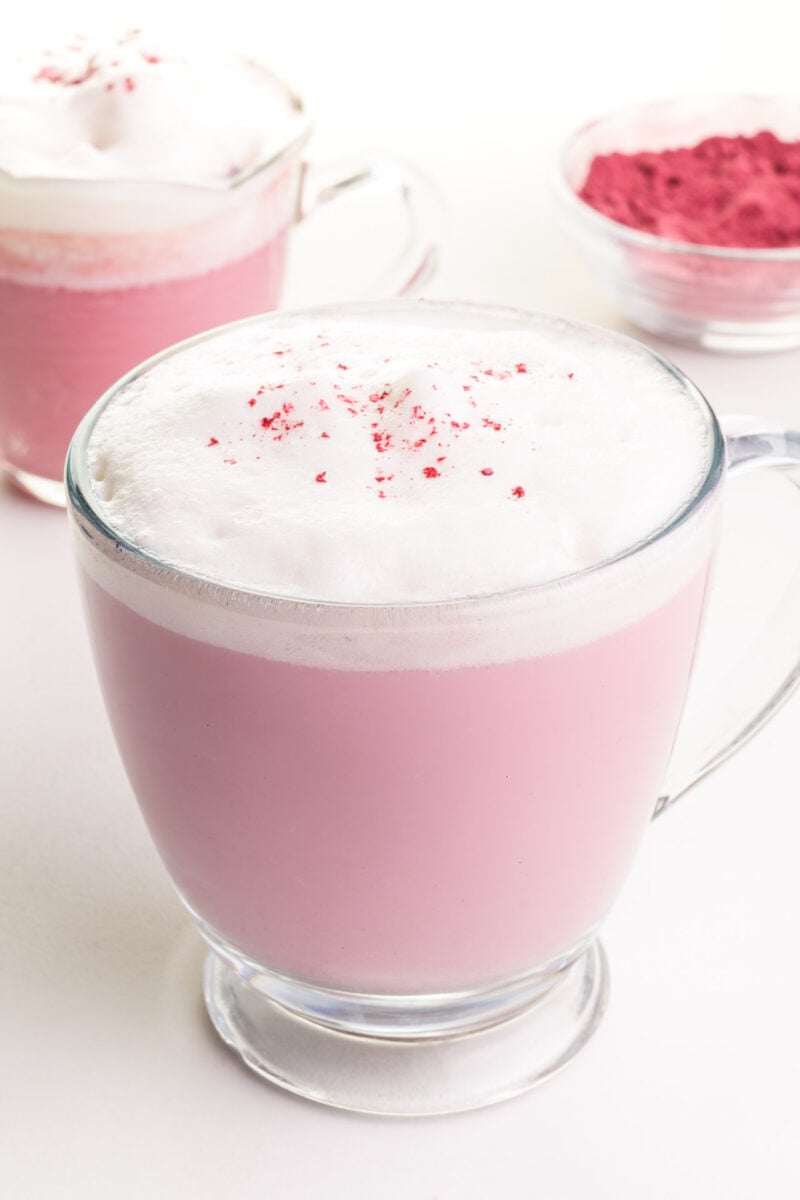A hot mug of pink matcha latte sits in front of another mug and the matcha powder in the background.