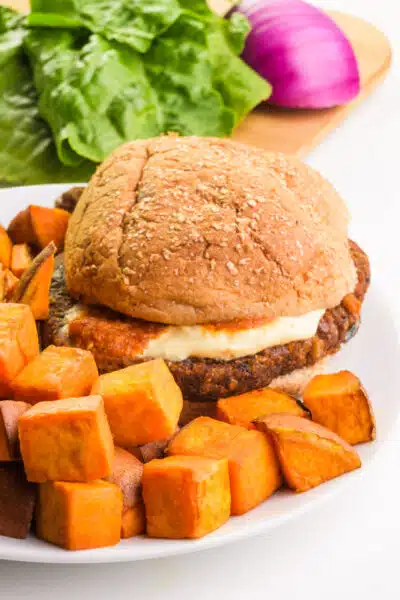 Sautéed sweet potatoes sit on a plate with a veggie burger. There are veggies and greens on a cutting board in the background.