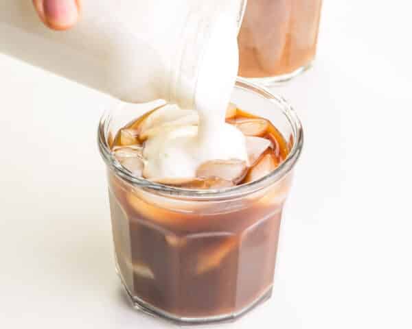Dairy-free cold foam is being poured over an iced coffee beverage.