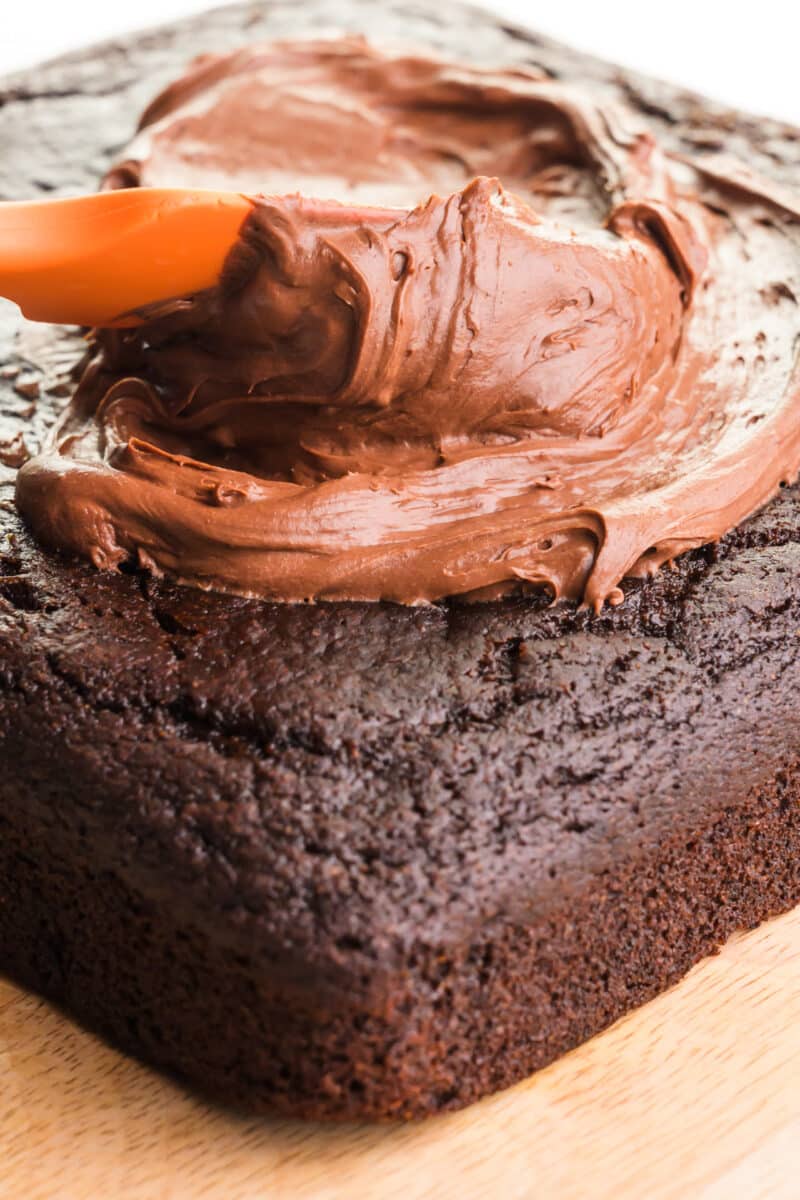 An engaging image portraying an orange spatula expertly spreading rich chocolate frosting on top of a freshly baked and wholesome healthy chocolate cake. The vibrant spatula contrasts with the indulgent chocolate frosting, creating a visually appealing scene of the final touch to this plant-based delight.