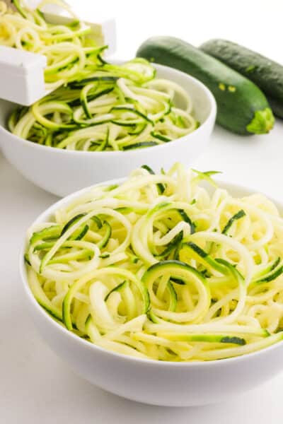 A bowl of zucchini noodles surrounded by fresh zucchini and a spiralizer creating more zoodles