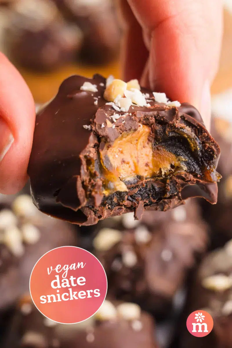 A hand holds a date bite with peanut butter in the center and dipped in chocolate. Text reads, vegan date snickers.