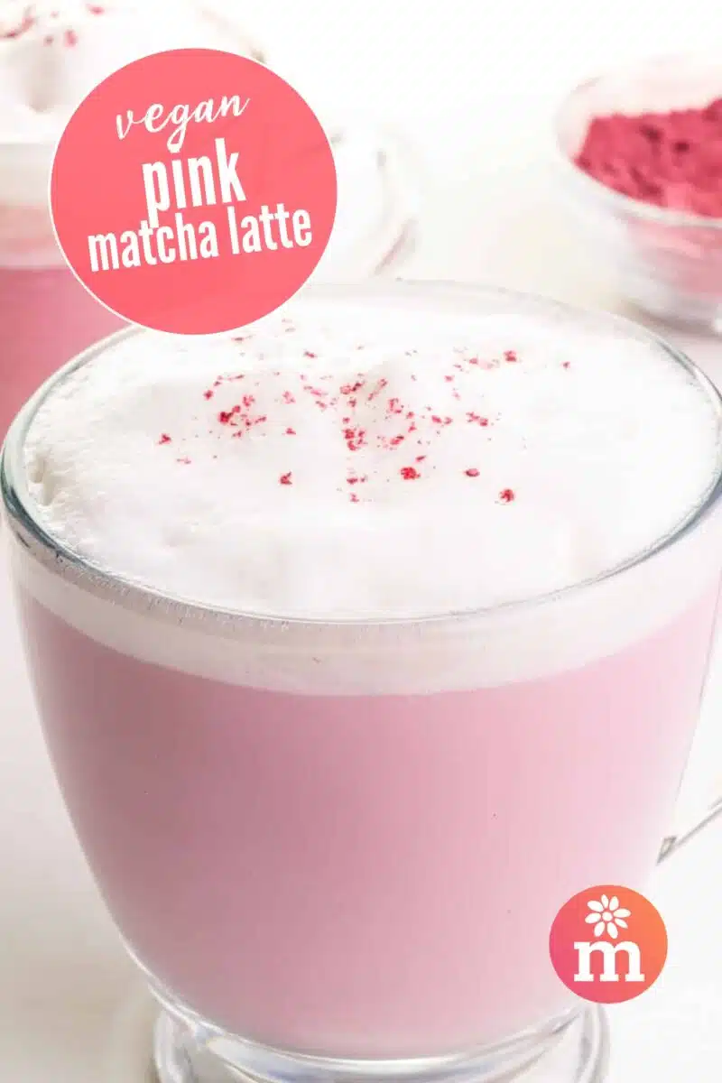 A closeup of a pink drink with white foam on top. Another mug and a bowl of pink matcha powder are in the background. Text reads: Vegan Pink Matcha Latte.