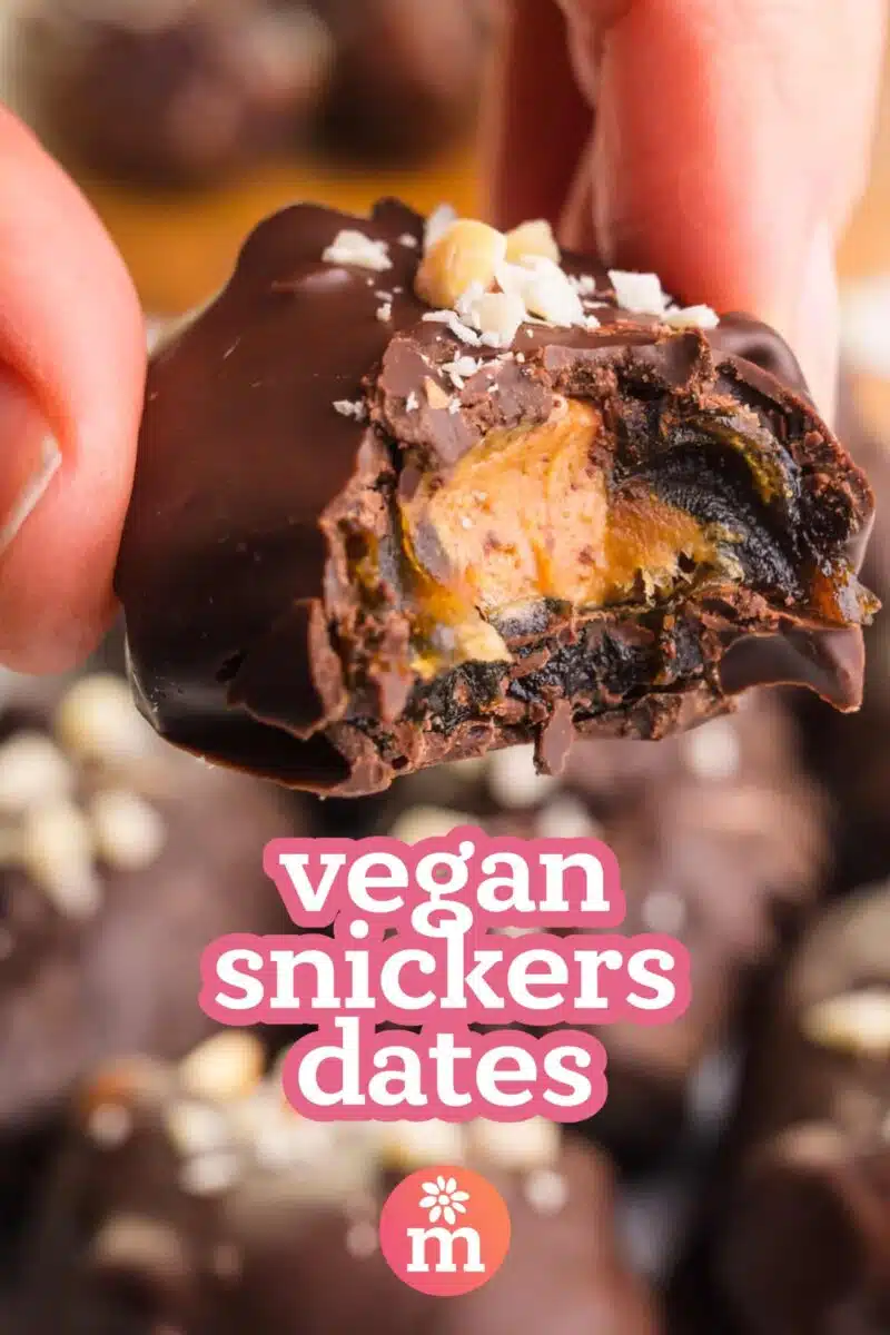 A date bite with peanut butter in the center and dipped in chocolate has a bite taken out. Text reads, vegan snickers dates.