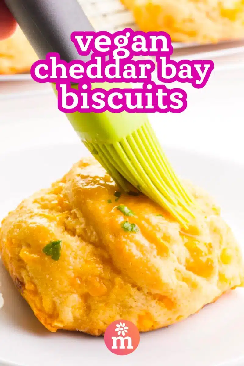 Melted butter is being brushed on a biscuit. The text reads, vegan cheddar bay biscuits.