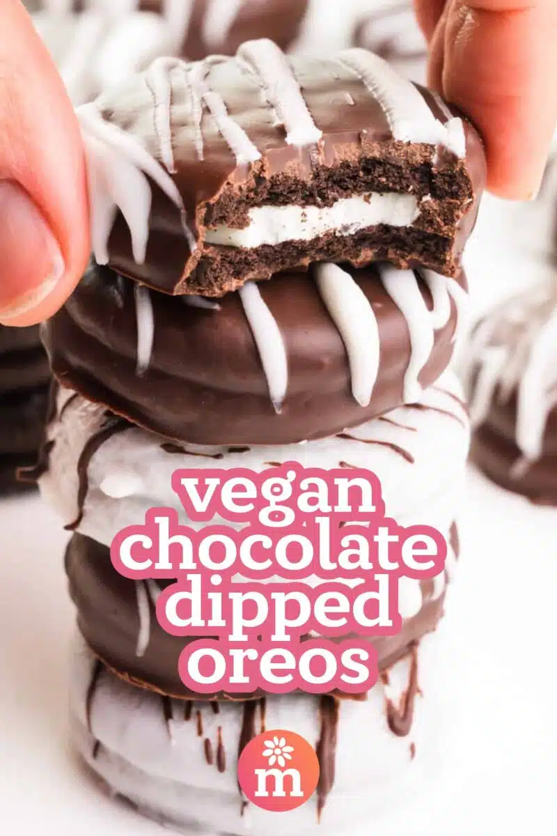 A hand grabs a cookie dipped in chocolate with a bite taken out. It sits on a stack of cookies. The text reads, vegan chocolate dipped Oreos.