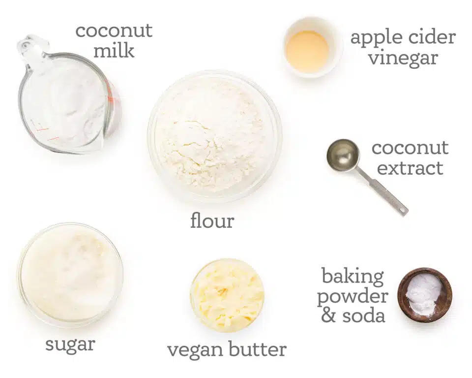 Ingredients are laid out on a white counter. The text next to them reads, apple cider vinegar, coconut extract, baking powder & soda, vegan butter, sugar, coconut milk, and flour.