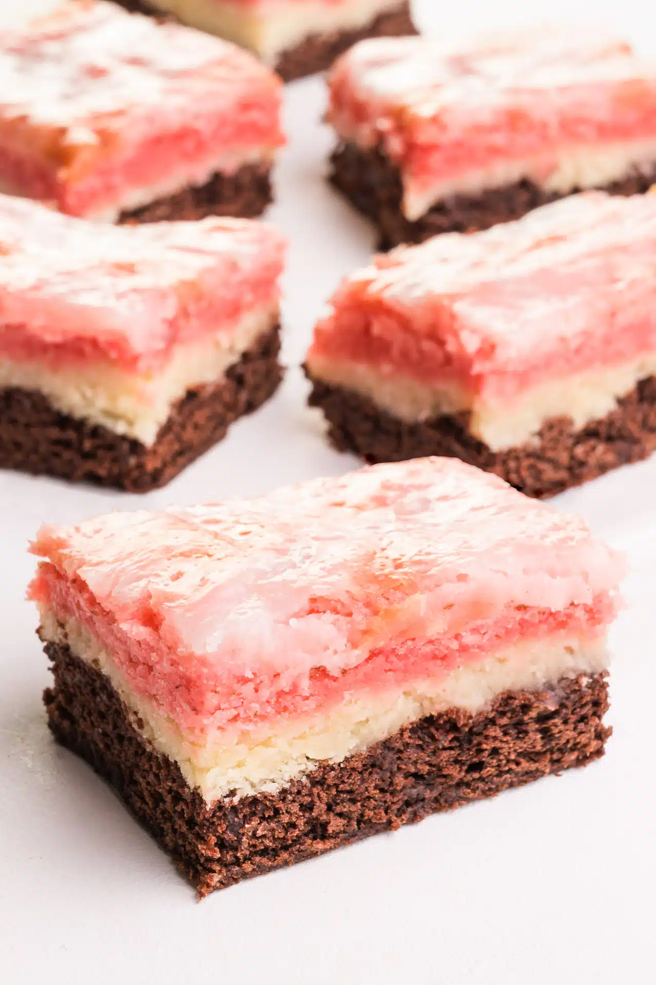 Several Neapolitan Bars sit on a white counter. The chocolate, vanilla, and strawberry treats treats have shiny tops from coconut sweetened condensed milk baked on top.