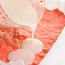 A coconut milk mixture is being poured into a pan with pink strawberry cake batter.