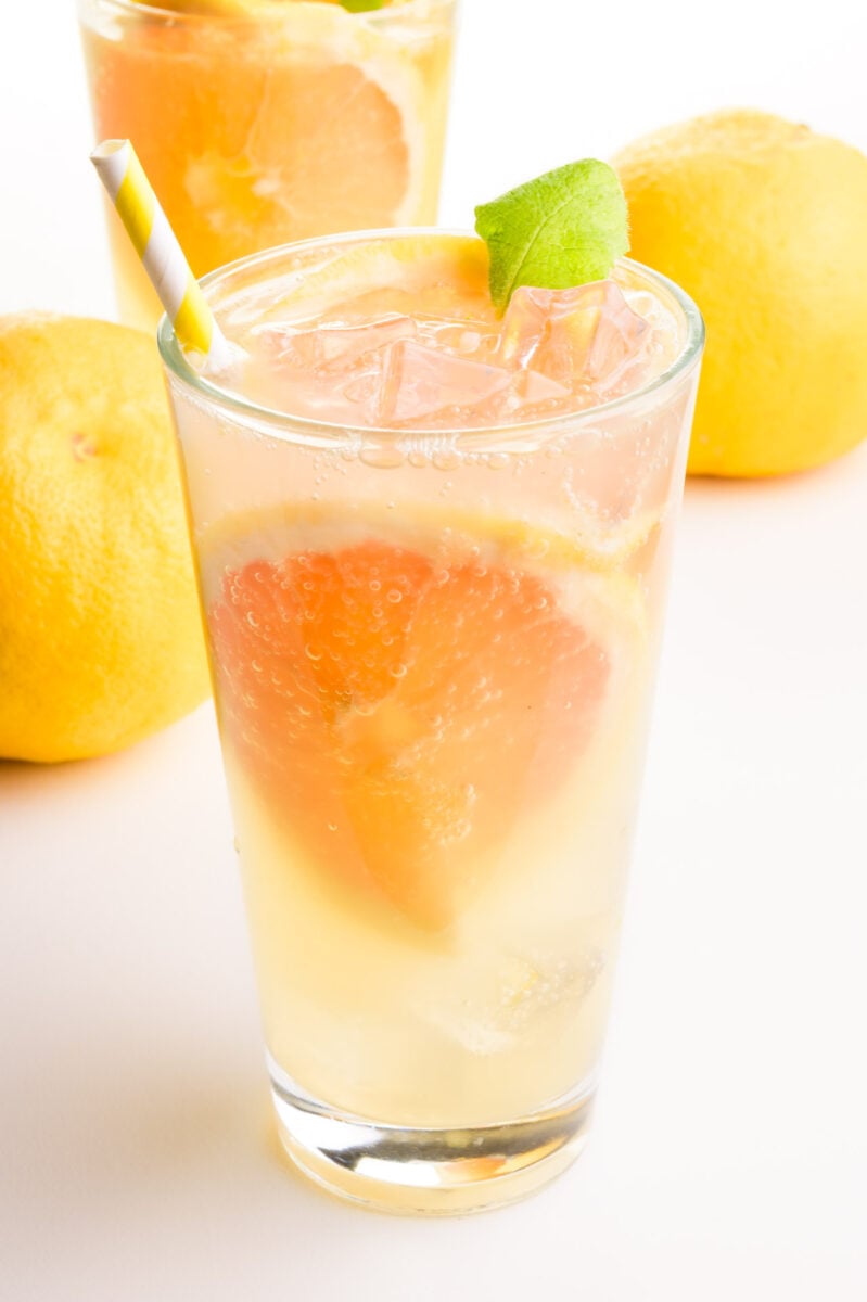 A glass of grapefruit green tea has grapefruit wedges in the glass. There are whole fruits in the background and another glass.