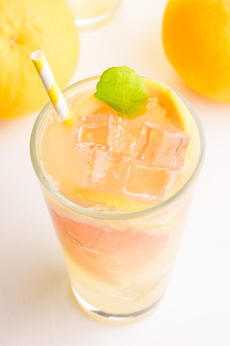 A glass holds a beverage with grapefruit a straw and a mint sprig. There are more grapefruits in the background.
