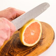A hand holds a knife, cutting a grapefruit on a cutting board.