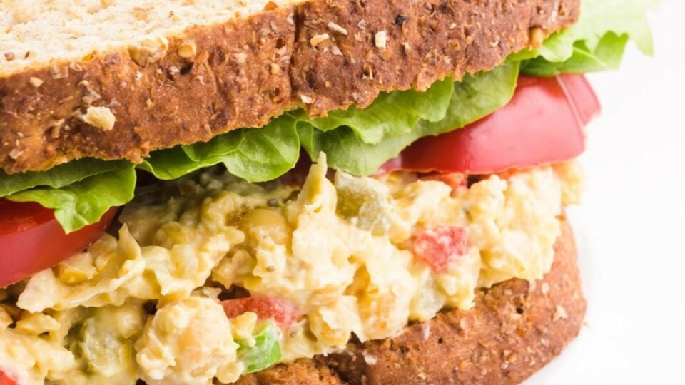 A closeup of a sandwich with chickpea salad topped with sliced veggies and lettuce leaves.