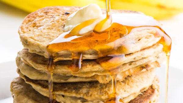 Maple syrup is being drizzled over banana pancakes with a pat of melty butter on top.