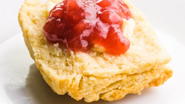 A closeup of a biscuit cut in half on a plate. It has butter and red jam on top.